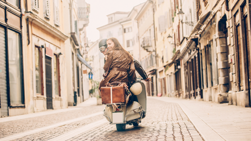 Dans une ruelle pavée, deux personnes sur un scooter vintage se retournent en souriant